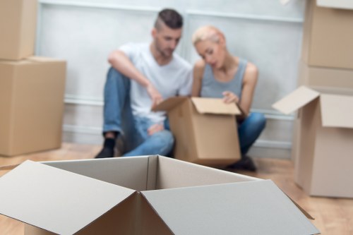 A man and a woman sit on the floor of a residential property in Lambeth, surrounded by multiple open and closed cardboard moving boxes of varying sizes, some showing flaps, on a light wooden floor. The man, with short dark hair and a beard, wears a casual light grey T-shirt and jeans, while the woman, with short platinum blonde hair, wears a grey sleeveless top. They are engaged in packing or unpacking, with the woman holding a small item from one of the boxes and the man appearing to assist or discuss the process. Behind them, a white wall and a staircase are visible, indicating they are in a hallway or living area in a typical London borough home. The scene is well-lit, with natural light coming from an unseen source, emphasizing the domestic environment and the activity of moving house, possibly in the Lambeth area, with the company Lambeth Man with Van providing removals services in the vicinity.