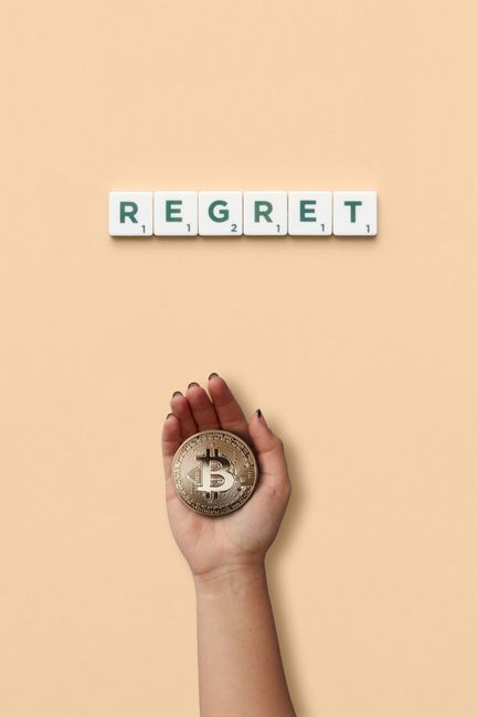 A hand holding a physical Bitcoin coin over a plain, light beige surface, with the word 'REGRET' spelled out in individual letter tiles arranged in a horizontal line above. The letter tiles are white 