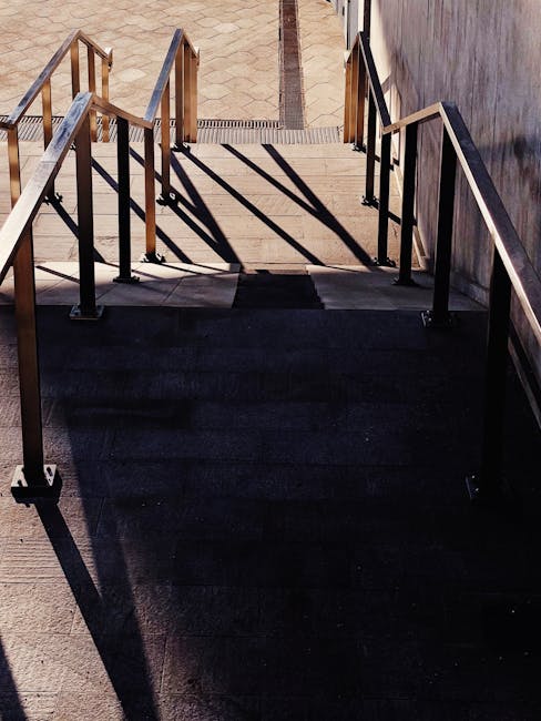 A downward view of a set of outdoor concrete stairs with metal handrails on both sides, leading from a paved sidewalk to a lower level outside a building. The stairs are partially shaded and the shado