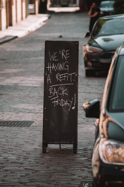A tall black chalkboard sign positioned on a cobblestone sidewalk, displaying handwritten white text that reads 'We're having a reffit back Friday!'. The sign is partially obscured by a parked vehicle